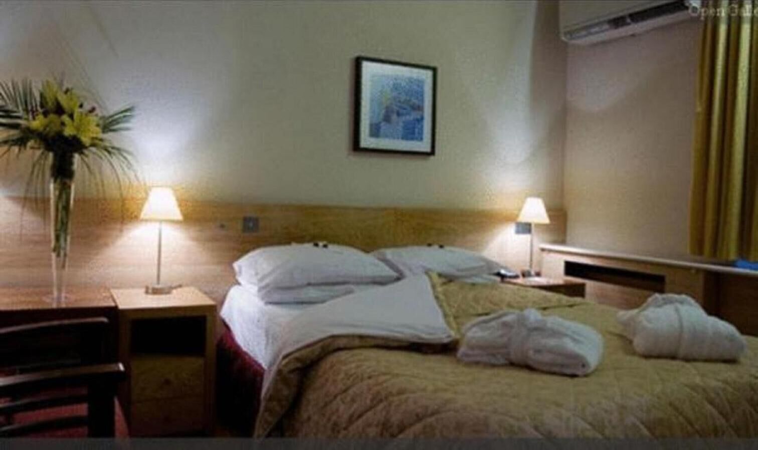 Habitación de hotel con cama, dos lámparas de noche, un escritorio, una silla, un jarrón con flores y un cuadro en la pared.