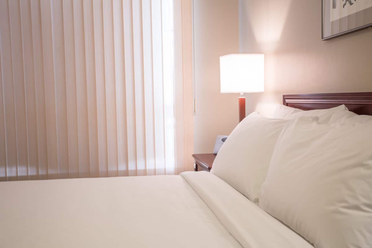 Ein Hotelzimmer mit einem ordentlich bezogenem Bett, einer Nachttischlampe und einem Fenster mit Jalousien.