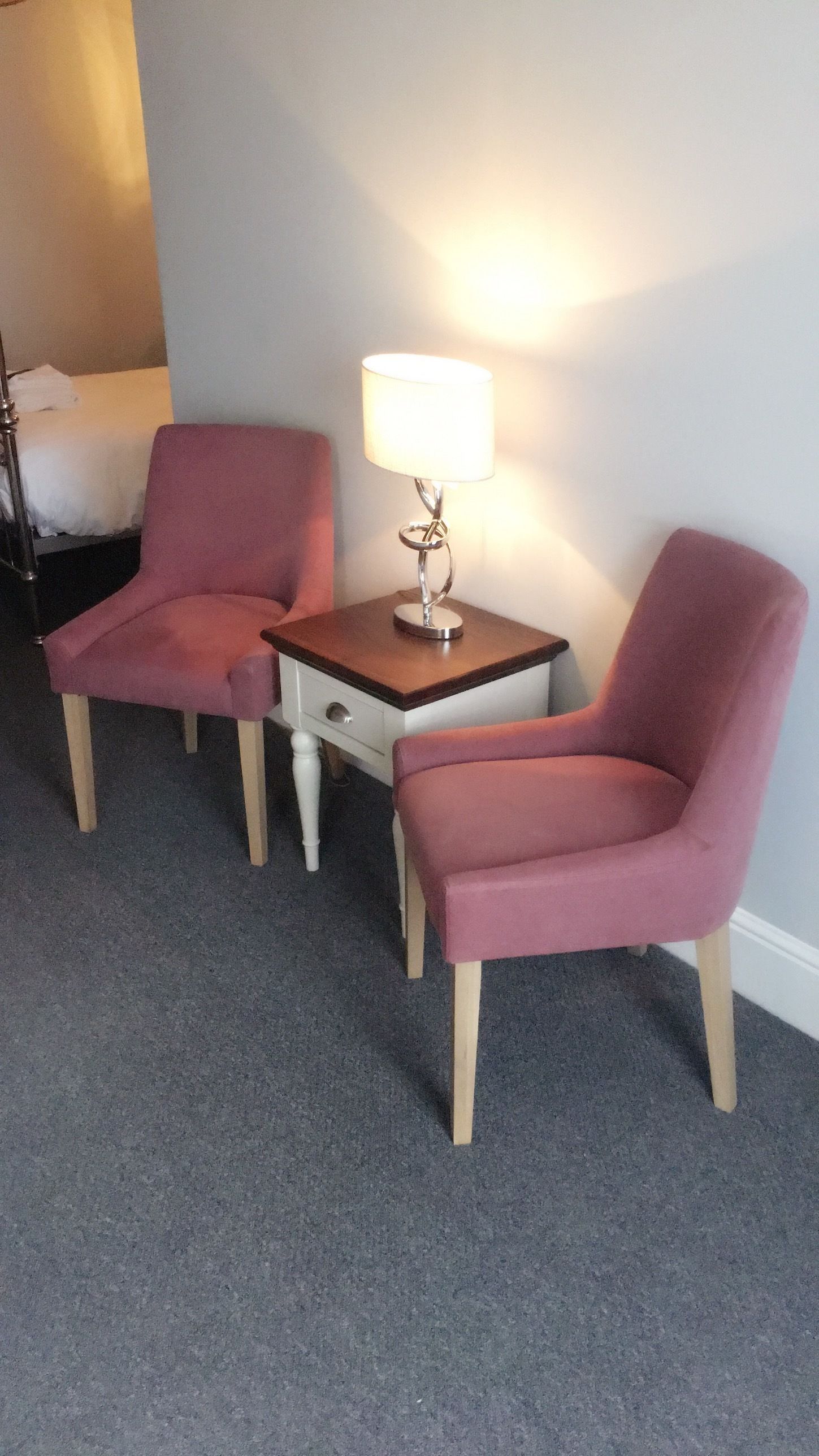 Habitación de hotel con dos sillones rosados, una pequeña mesa de madera con una lámpara y una cama al fondo.
