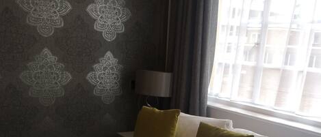 A hotel room with a bed, a window with curtains, a lamp, and a wall with a floral pattern.