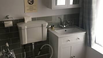 Ein Badezimmer mit Toilette, Waschbecken und Badewanne. An der Wand ist ein Warnschild angebracht, und es gibt ein Fenster mit Vorhängen.