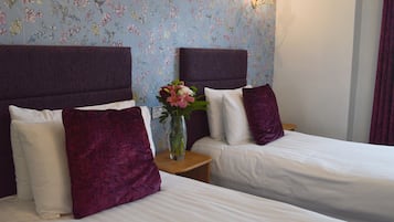 Habitación de hotel con dos camas, cada una con cabecera, ropa de cama blanca y almohada morada. Entre las camas hay una mesita con un florero.