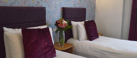 Habitación de hotel con dos camas, cada una con cabecera, ropa de cama blanca y almohada morada. Entre las camas hay una mesita con un florero.