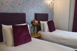 Habitación de hotel con dos camas, cada una con cabecera, ropa de cama blanca y almohada morada. Entre las camas hay una mesita con un florero.