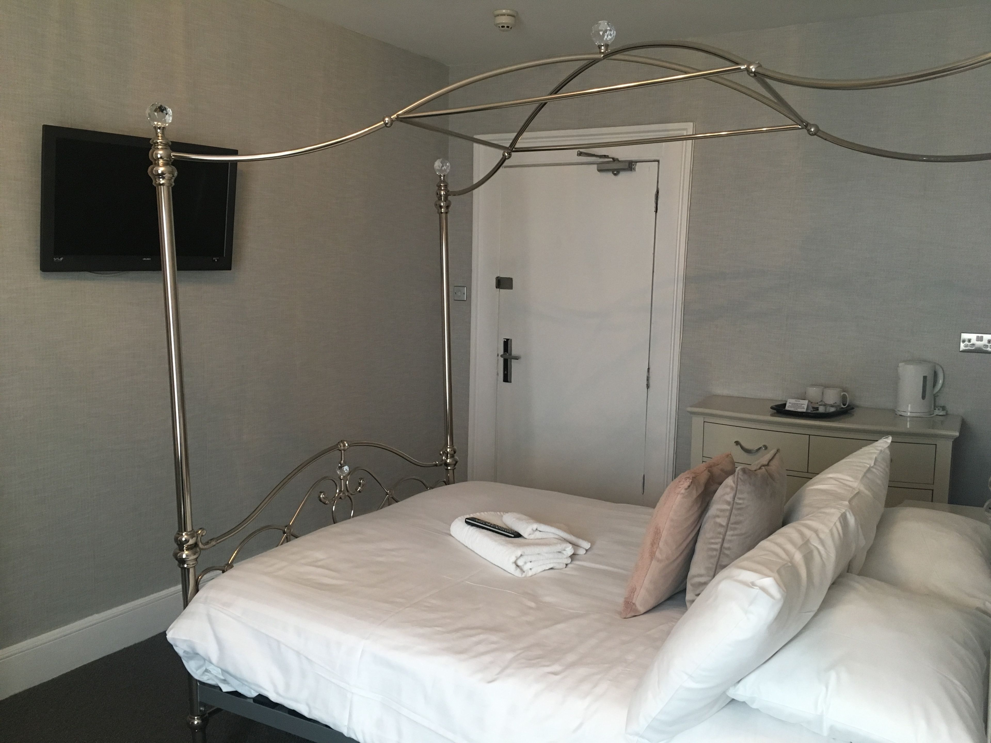A neatly made bed with white linens, a bedside table with a cup and a tray of cups, and a flat-screen TV mounted on the wall.