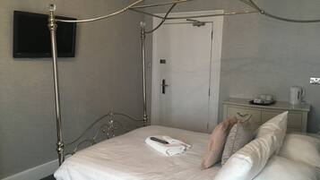A neatly made bed with white linens, a bedside table with a cup and a tray of cups, and a flat-screen TV mounted on the wall.