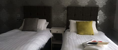 A hotel room with two beds, each with white bedding and a headboard. There is a nightstand between the beds with a lamp and a remote control on it. The walls have a patterned wallpaper.