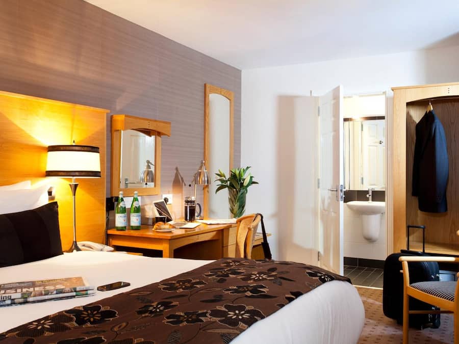 A hotel room with a bed, bedside table, lamp, mirror, desk, chair, and a wardrobe.