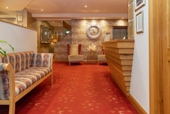 Un hall de hotel con alfombra roja, un sofá, dos sillones, un mostrador de recepción y un reloj de pared.