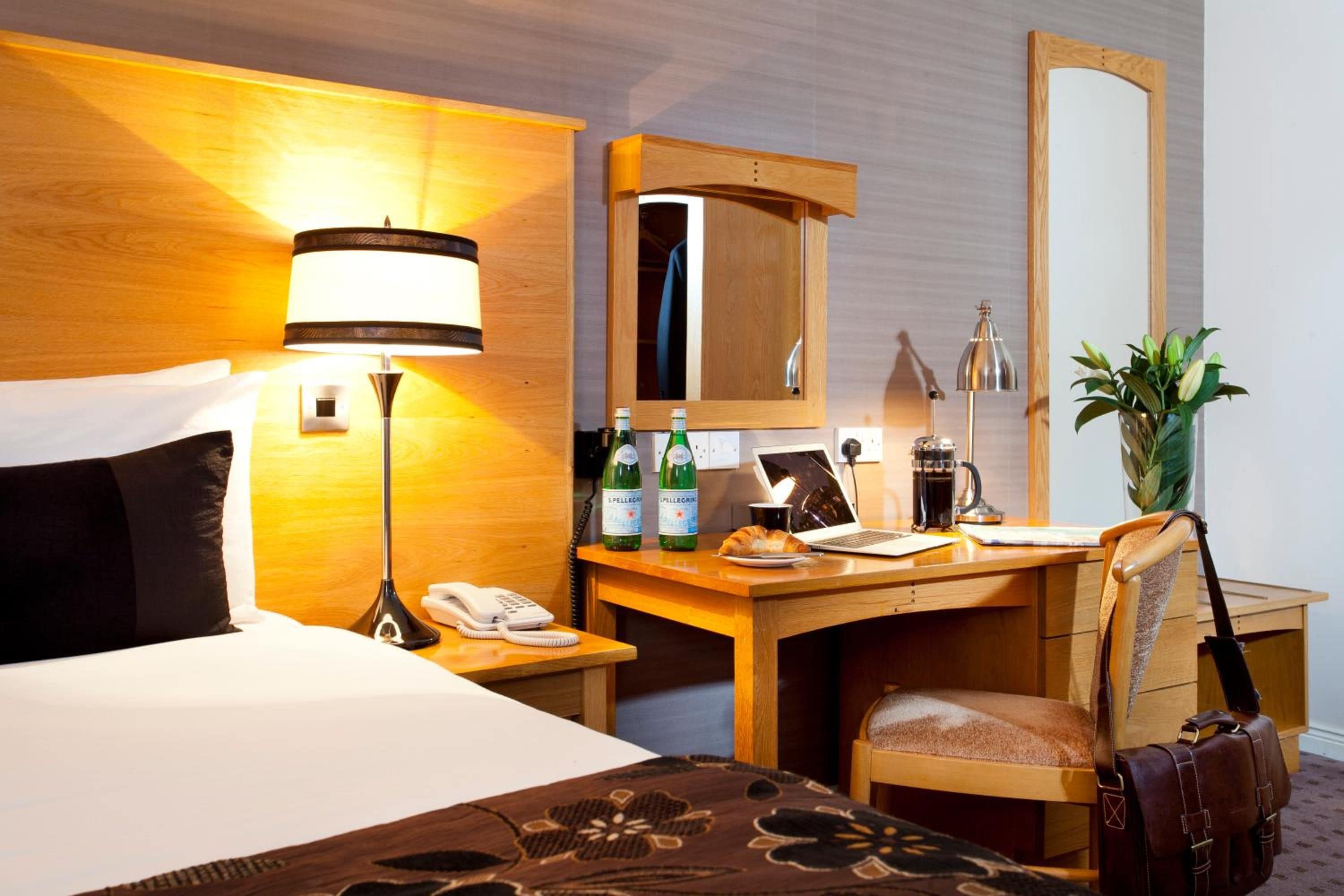 Habitación de hotel con cama, escritorio, silla, lámpara, espejo y planta en maceta.