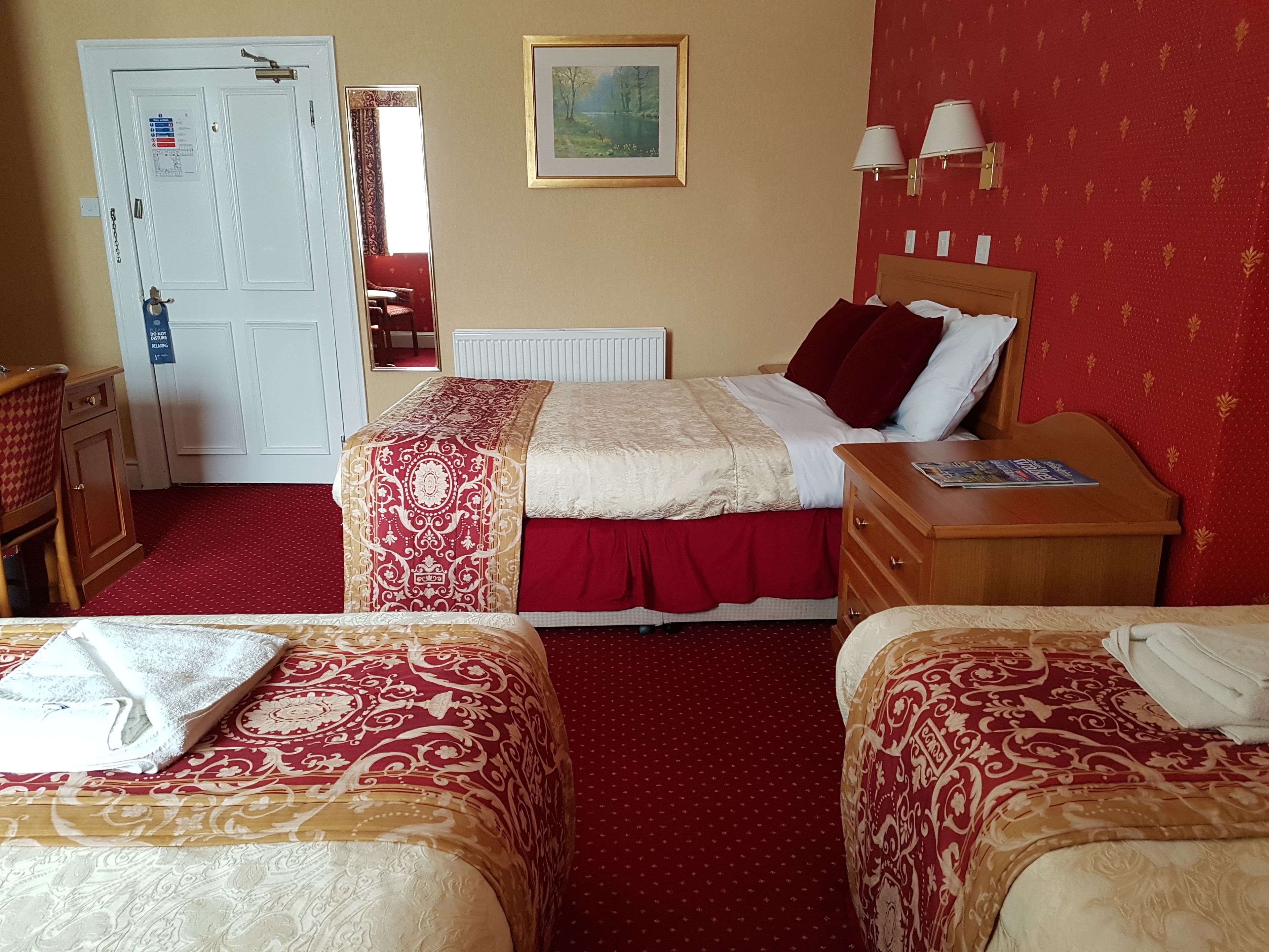Une chambre d’hôtel avec deux lits, une table de chevet en bois, une commode, une moquette rouge et un tableau au mur.