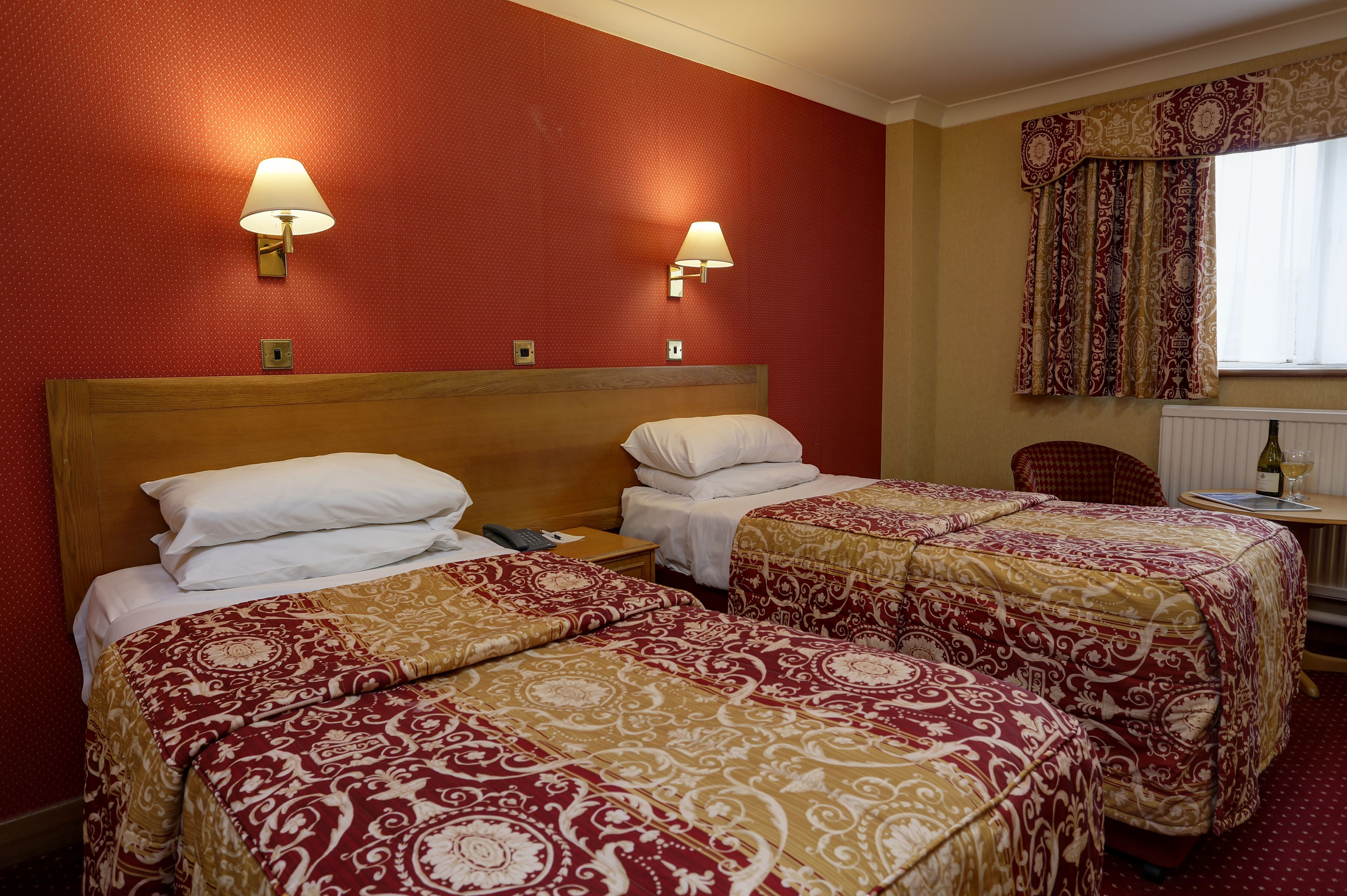 A hotel room with two beds, a wooden headboard, a small table with a bottle and glass, and two wall-mounted lamps.