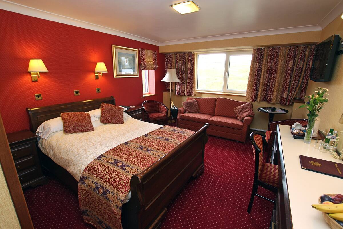 A hotel room with a bed, two chairs, a table with a fruit bowl, a TV, and a window with curtains.
