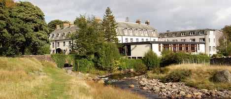 A riverside hotel with a stone pathway leading to the entrance, surrounded by lush greenery and a calm river.