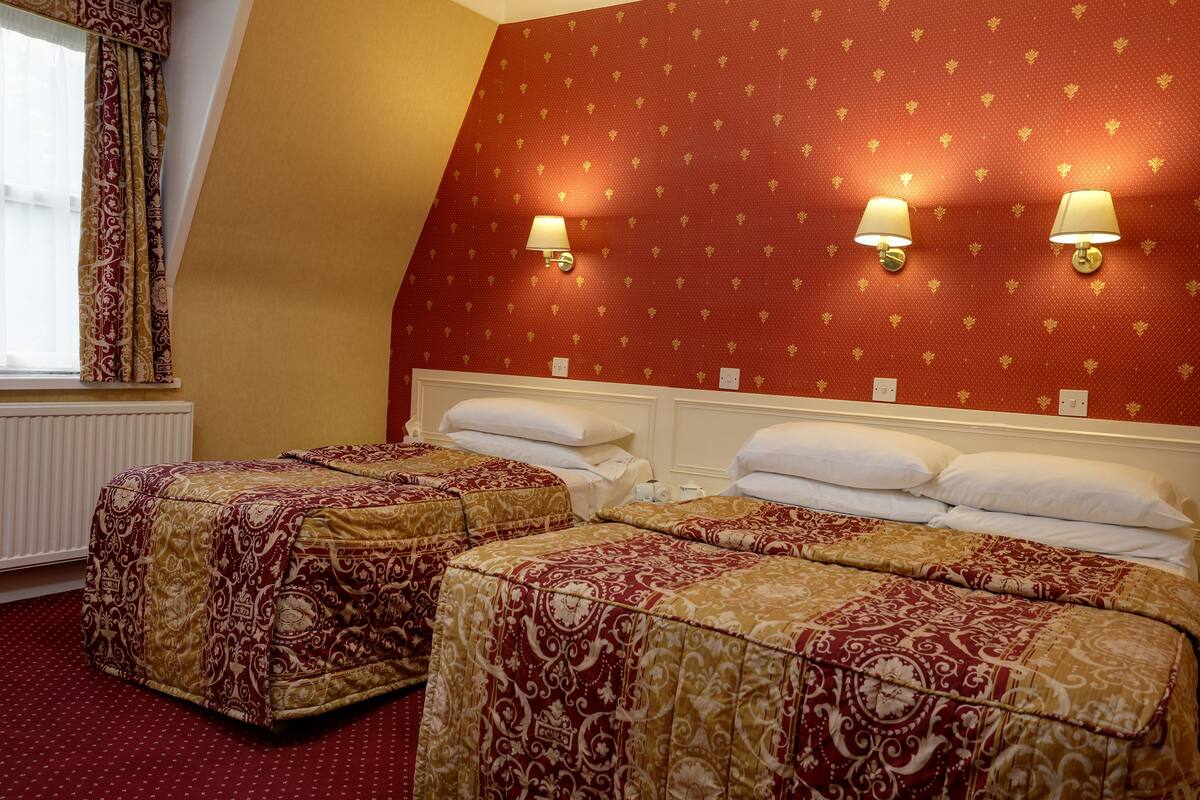 A hotel room with two beds, red patterned wallpaper, and two wall-mounted lamps.