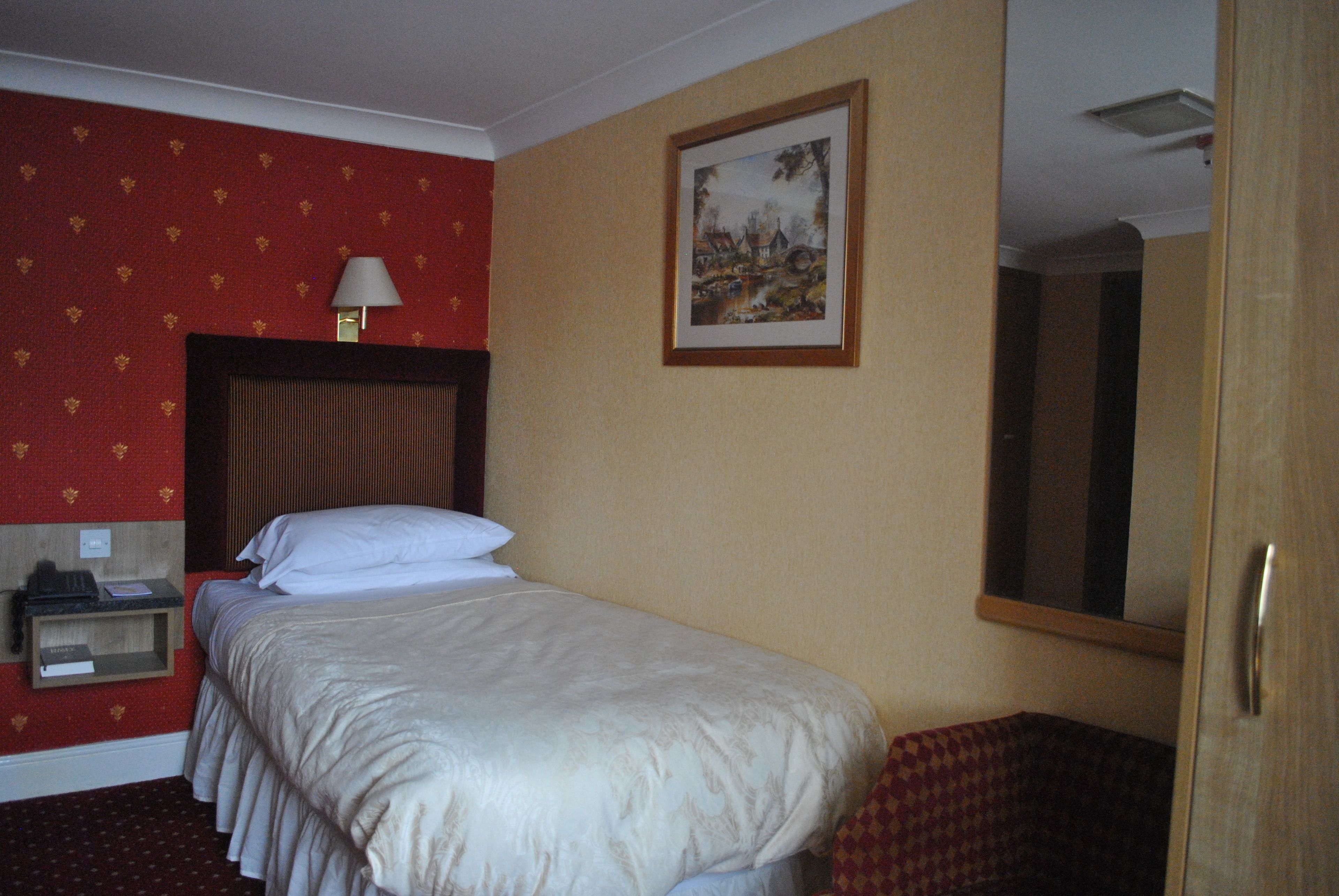 Quarto de hotel com cama, mesa de cabeceira, abajur, quadro na parede e porta.