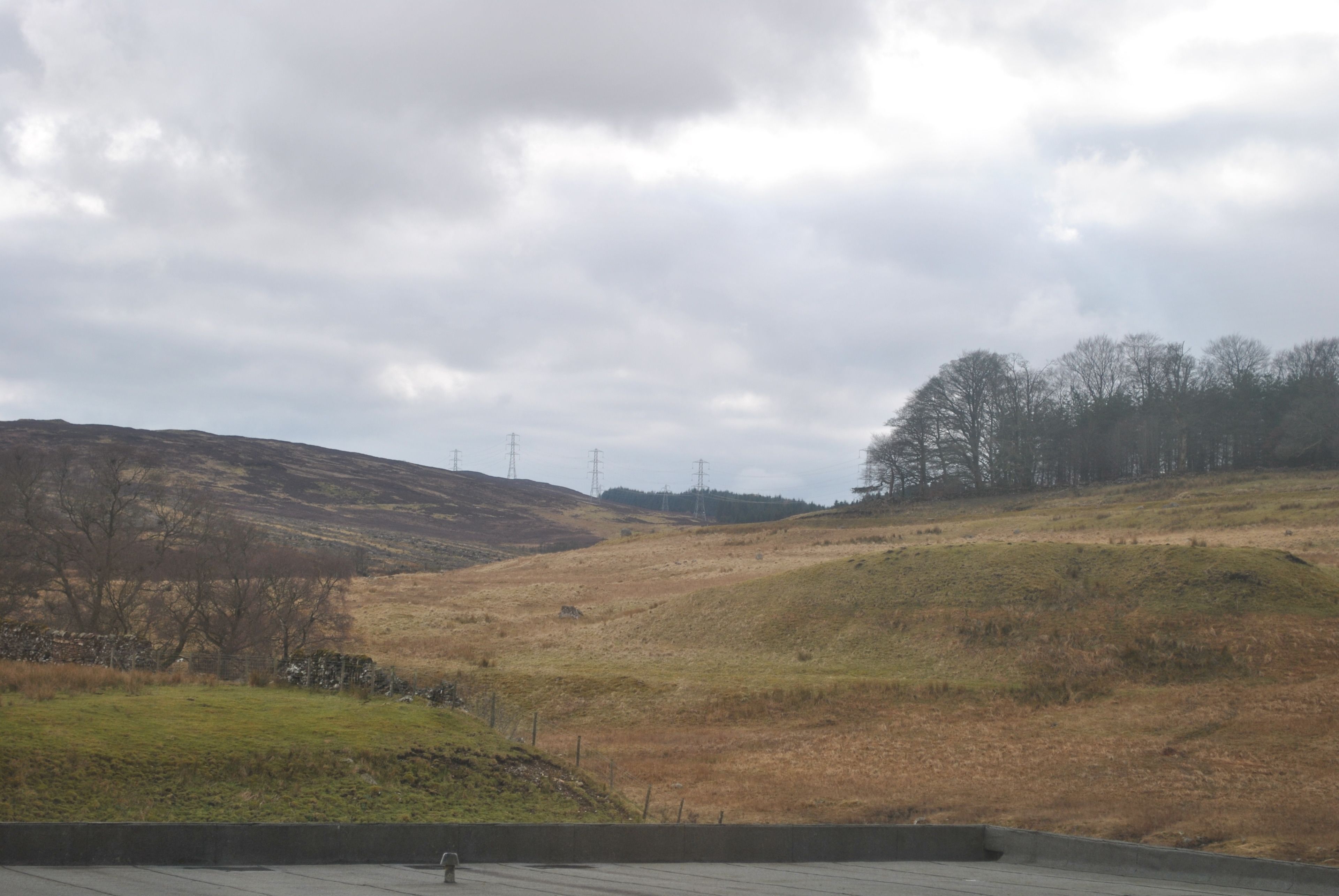 A rural landscape with a grassy hill, a stone wall, and power lines in the distance.