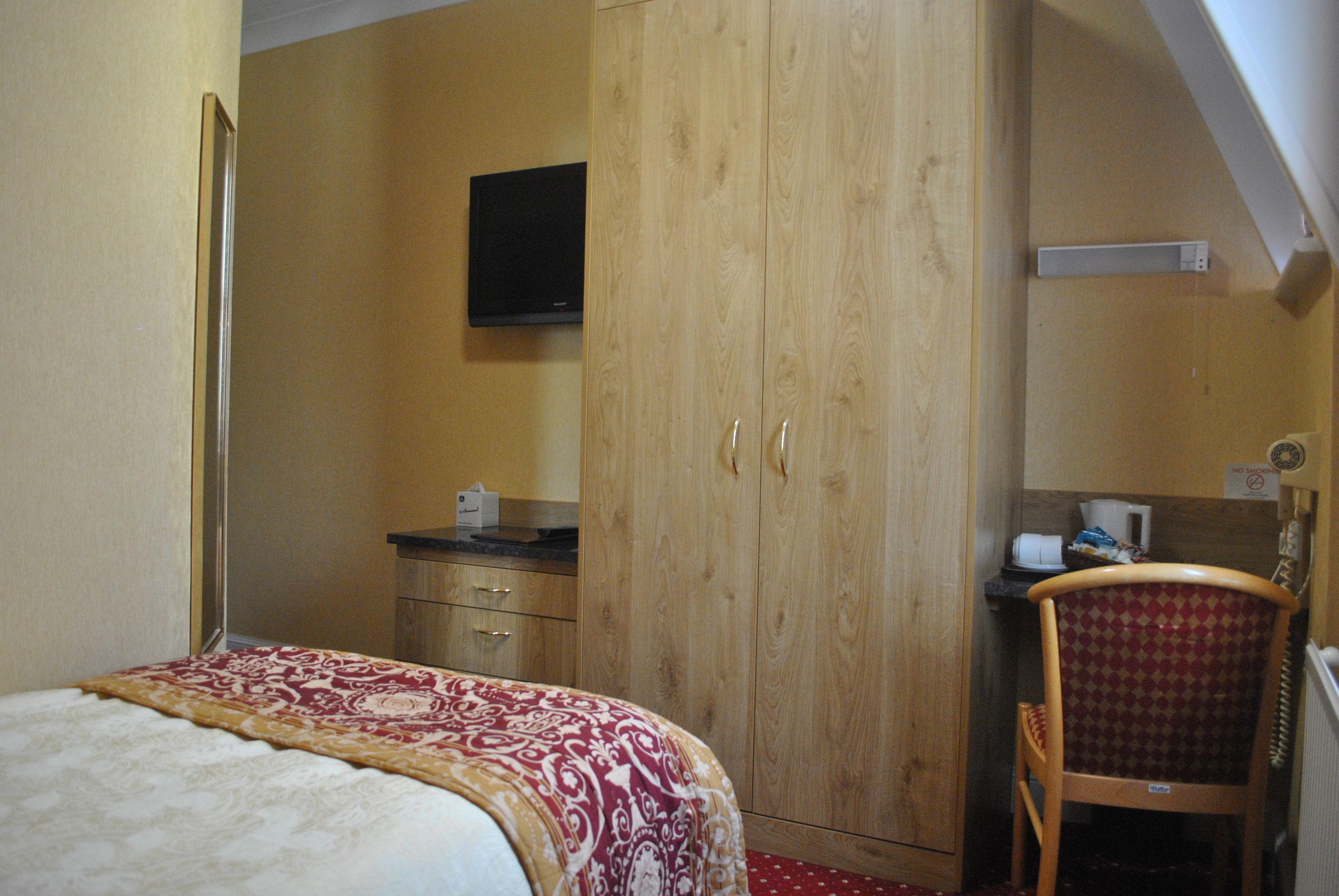 Une chambre à coucher avec un lit, une armoire en bois, une télévision, une chaise et une petite table sur laquelle se trouvent une tasse et des mouchoirs.