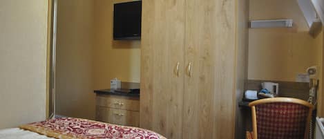 A bedroom with a bed, wooden wardrobe, a television, a chair, and a small table with a cup and tissues.