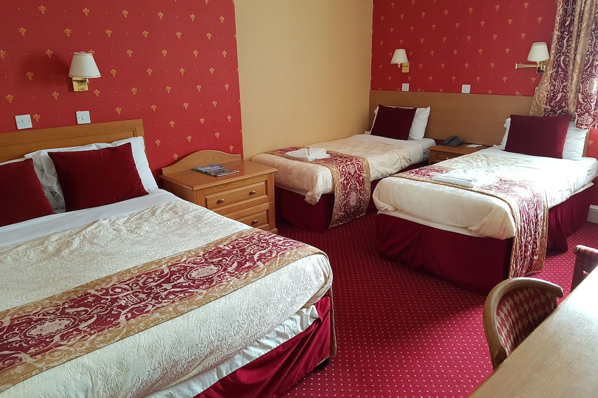 A hotel room with two beds, each with red and white bedding, a wooden nightstand, a small table, and a red carpeted floor.