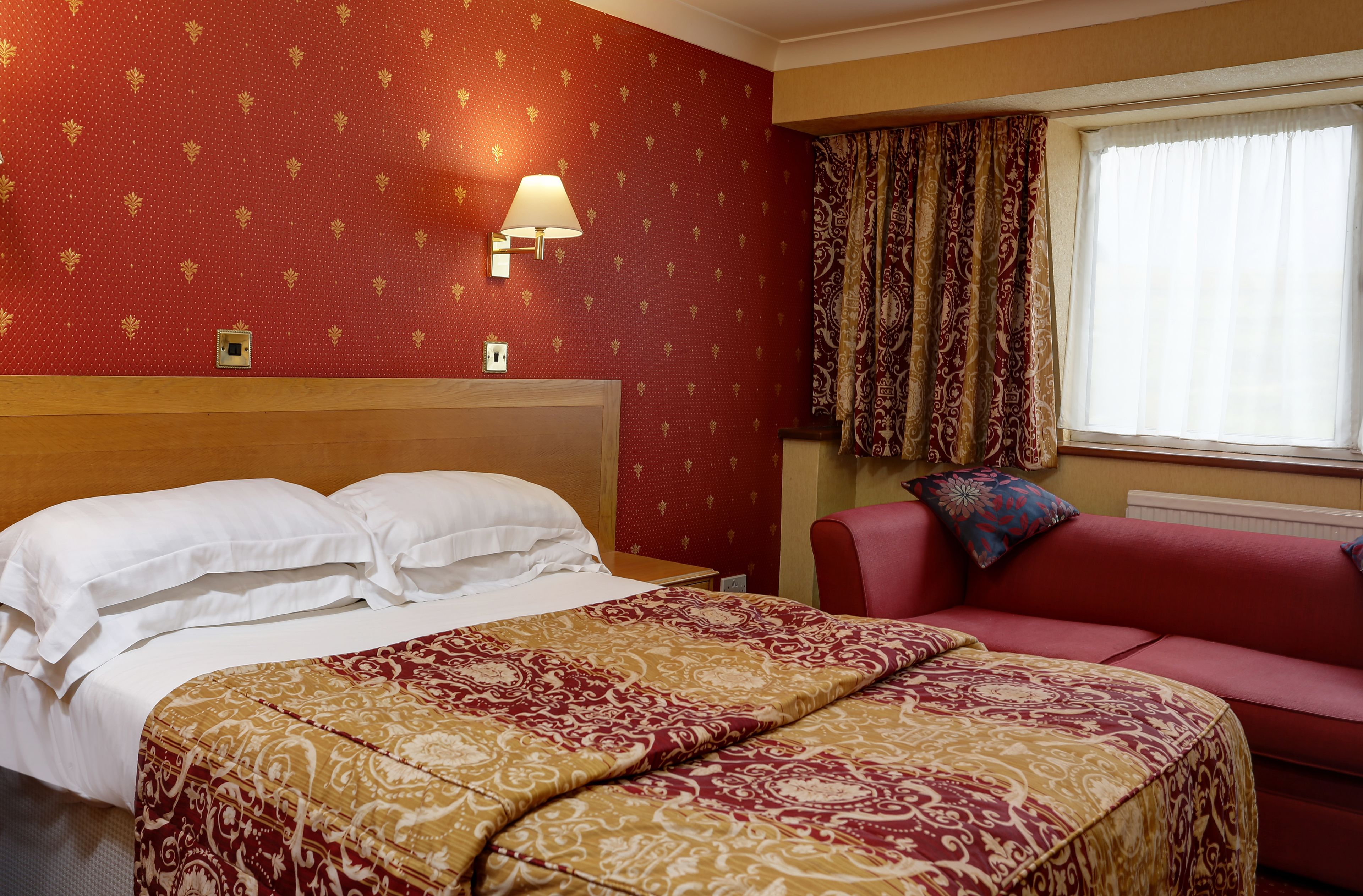 Una habitación de hotel con una cama, un sofá rojo, una manta estampada, una lámpara de pared y una ventana con cortinas.