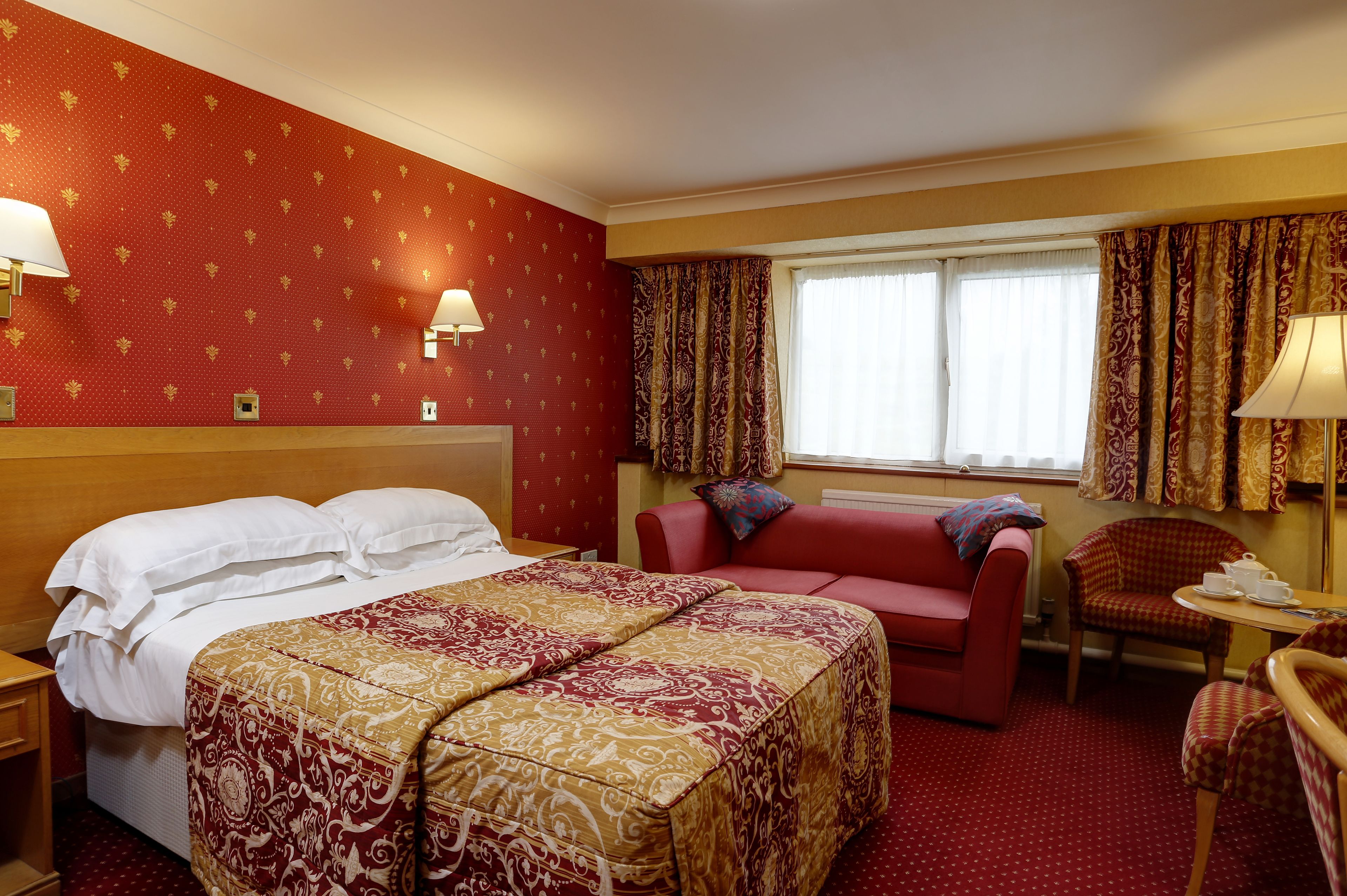 Ein Hotelzimmer mit einem großen Bett, einem roten Sofa, einem roten Sessel und einem kleinen Tisch mit einem Teekessel.