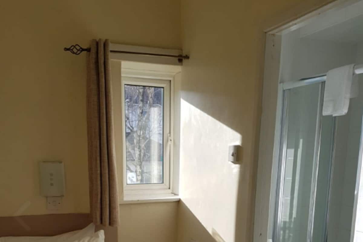 Une chambre d’hôtel comprenant un lit, une petite table de chevet, une fenêtre avec des rideaux, un espace douche doté d’une porte en verre et un sol carrelé.