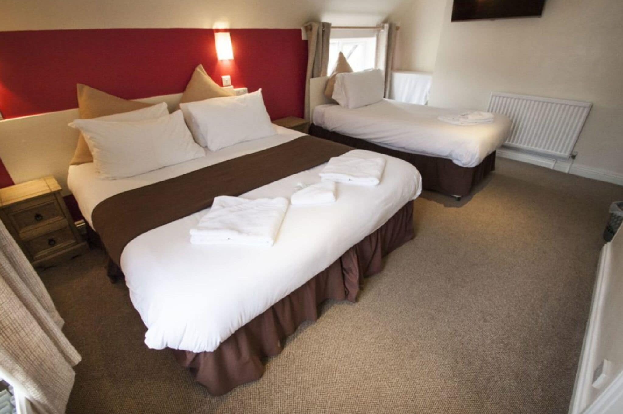 A hotel room with two beds, a red headboard, a white wall, and a window with curtains.