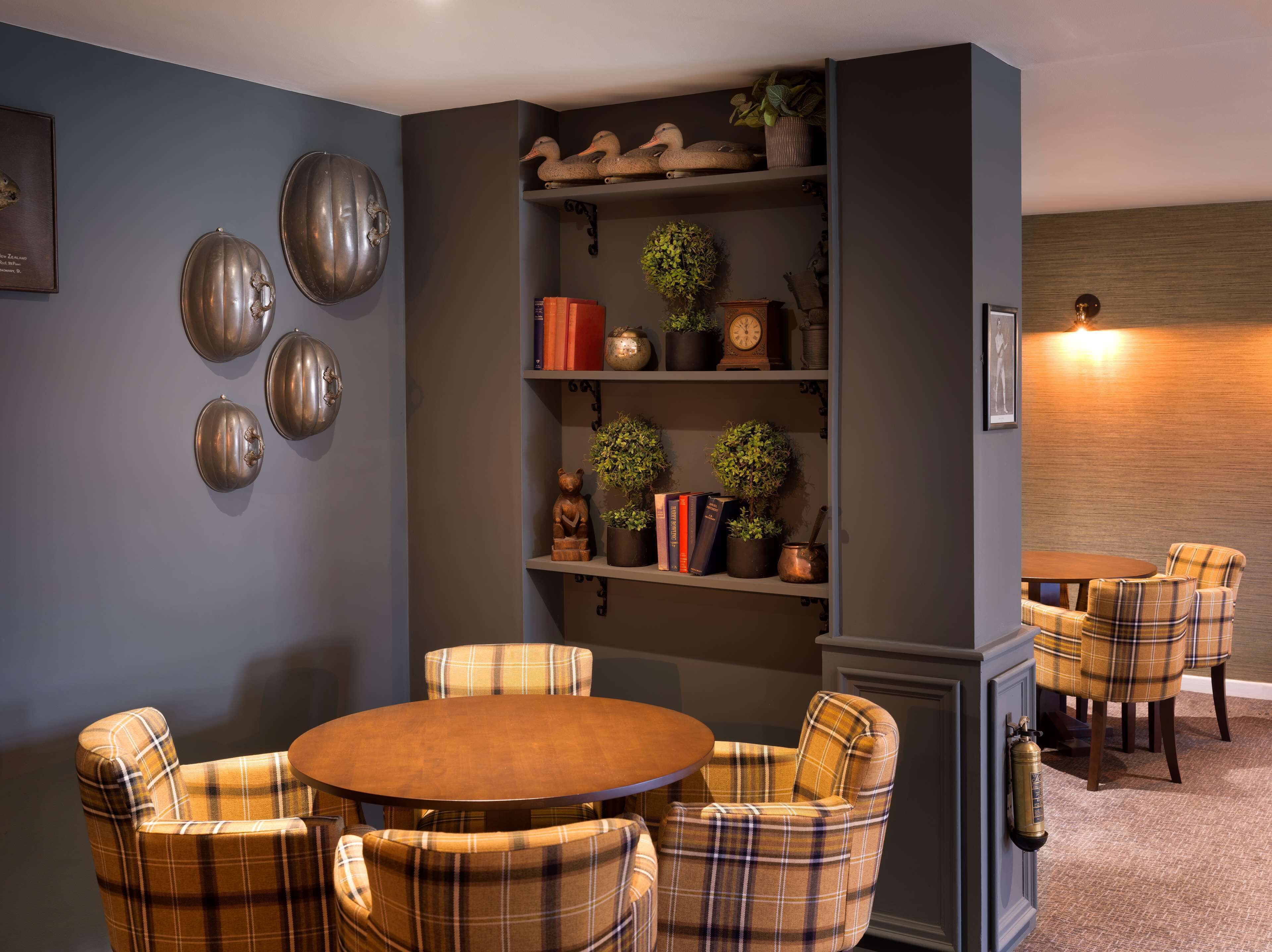 A dining area with a round table and plaid chairs, a bookshelf with decorative items, and a wall with a clock and wall art.