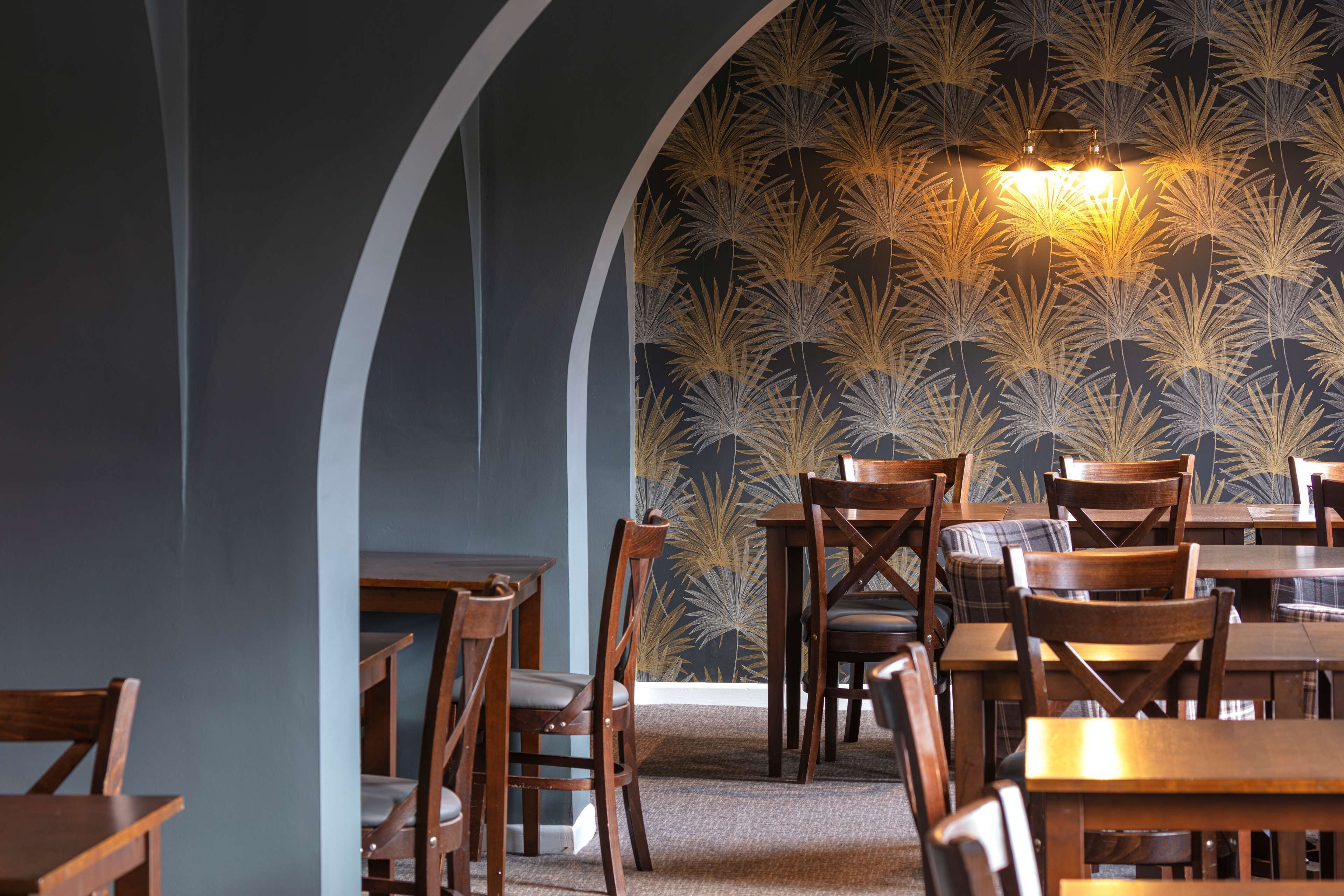 Ein Restaurantinneres mit Holztischen und -stühlen, einem gemusterten Wandpapier und einem außergewöhnlichen Deckenstil.