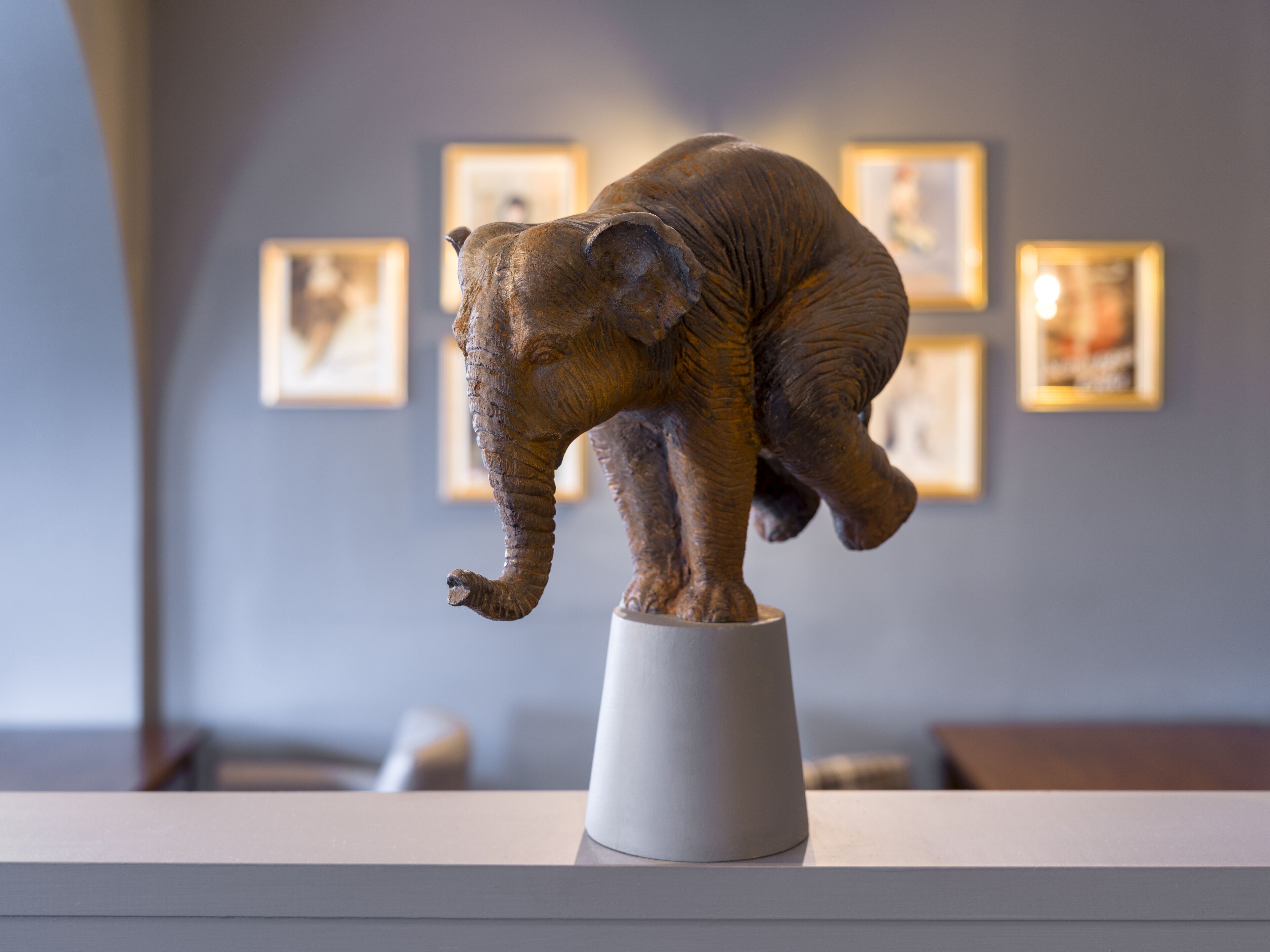 A sculpture of an elephant on a pedestal in a room with framed pictures on the wall.