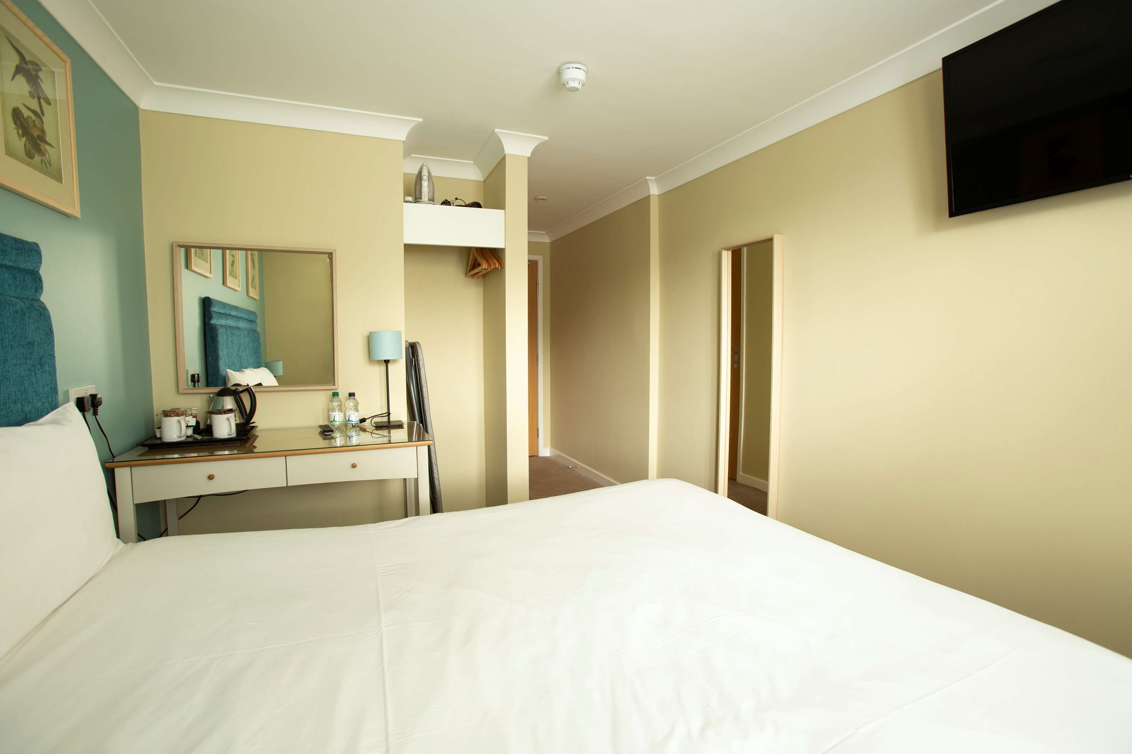 A hotel room with a bed, a desk with a mirror, a flat-screen TV, and a door leading to another room.