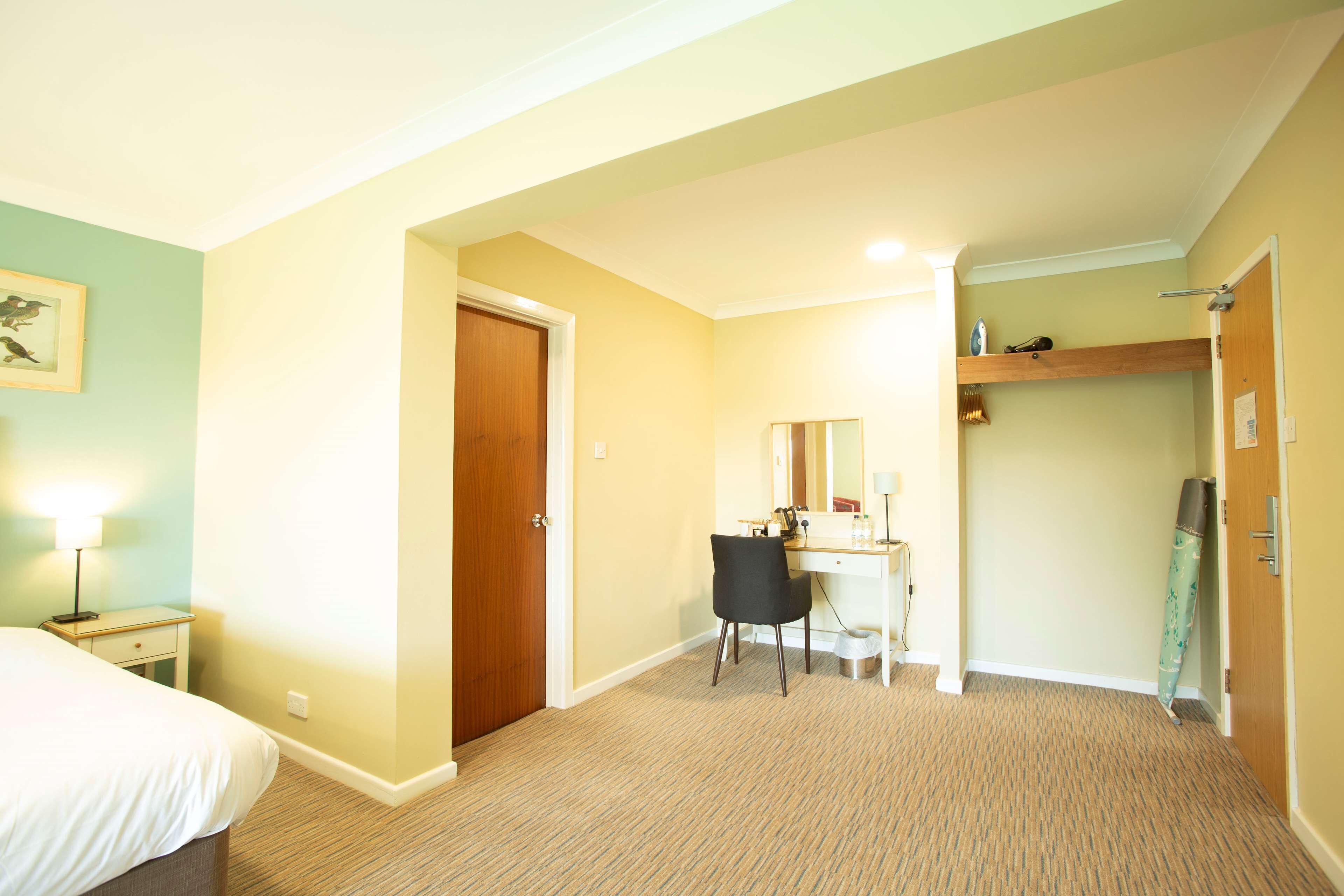 A hotel room with a bed, a desk, a chair, a mirror, a shelf with toiletries, and a door.