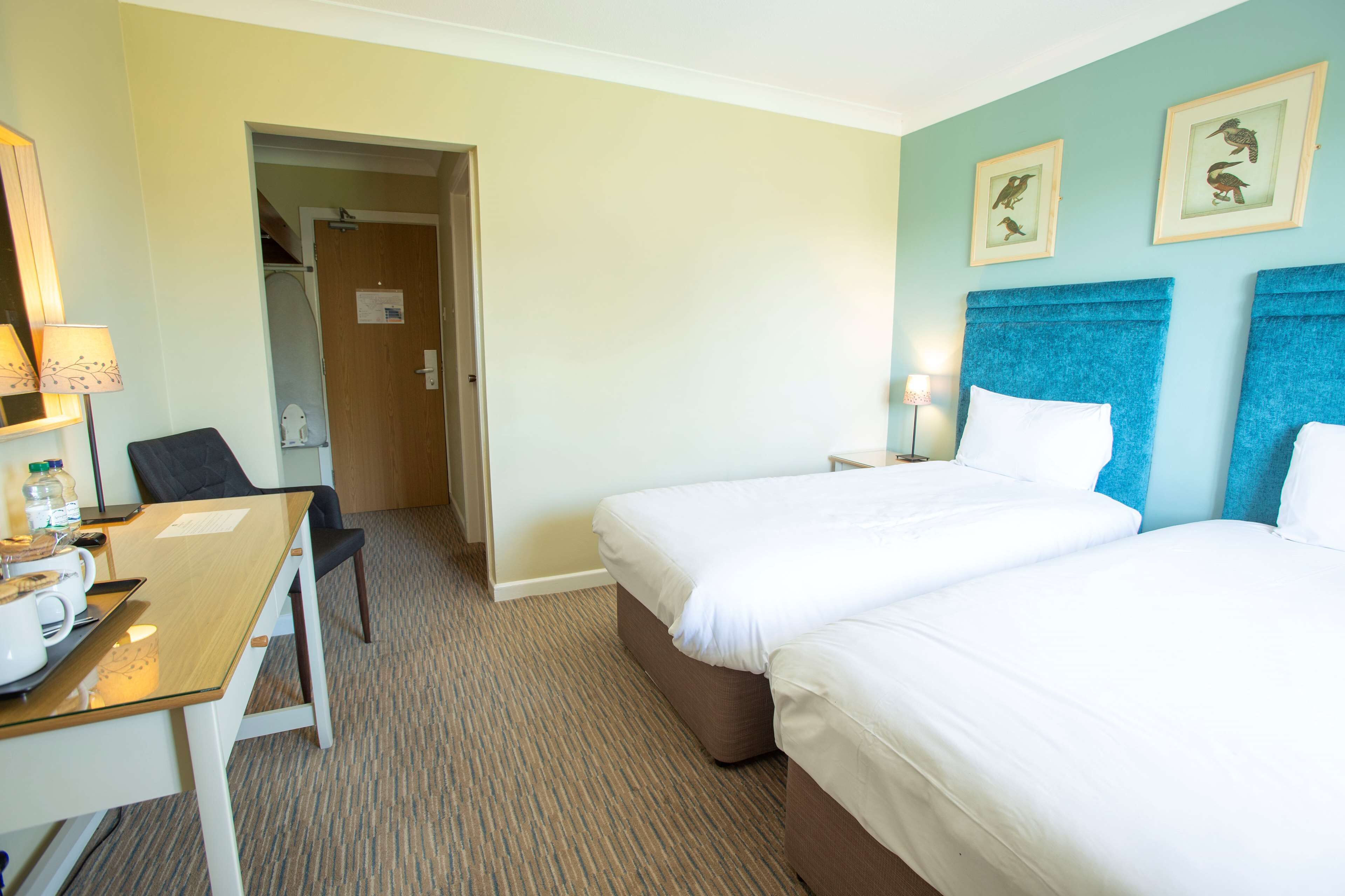 A hotel room with two beds, a desk with a chair, a lamp, and a door leading to a bathroom.