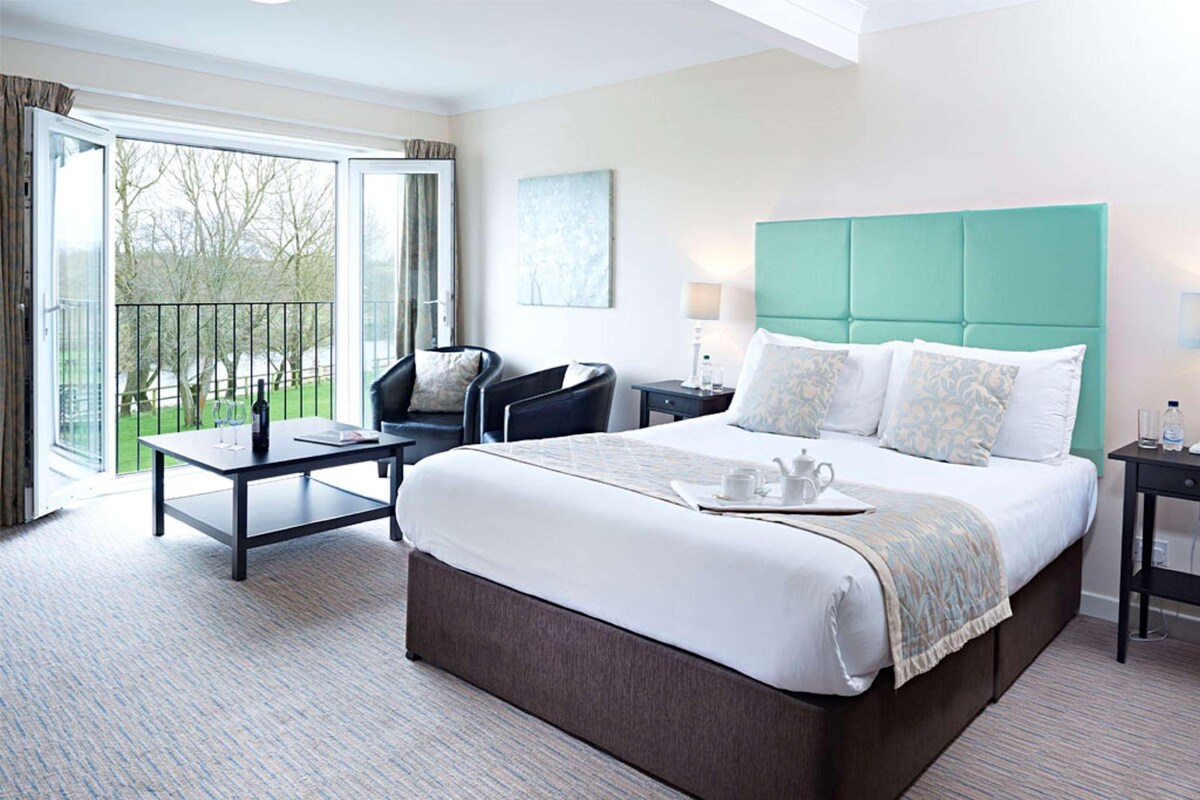 A hotel room with a large bed, a desk, a chair, and a balcony with a view of trees.
