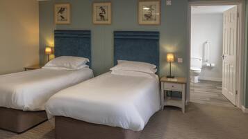 Habitación de hotel con dos camas, mesitas de noche, lámparas, cuadros en la pared y un baño visible a través de una puerta abierta.