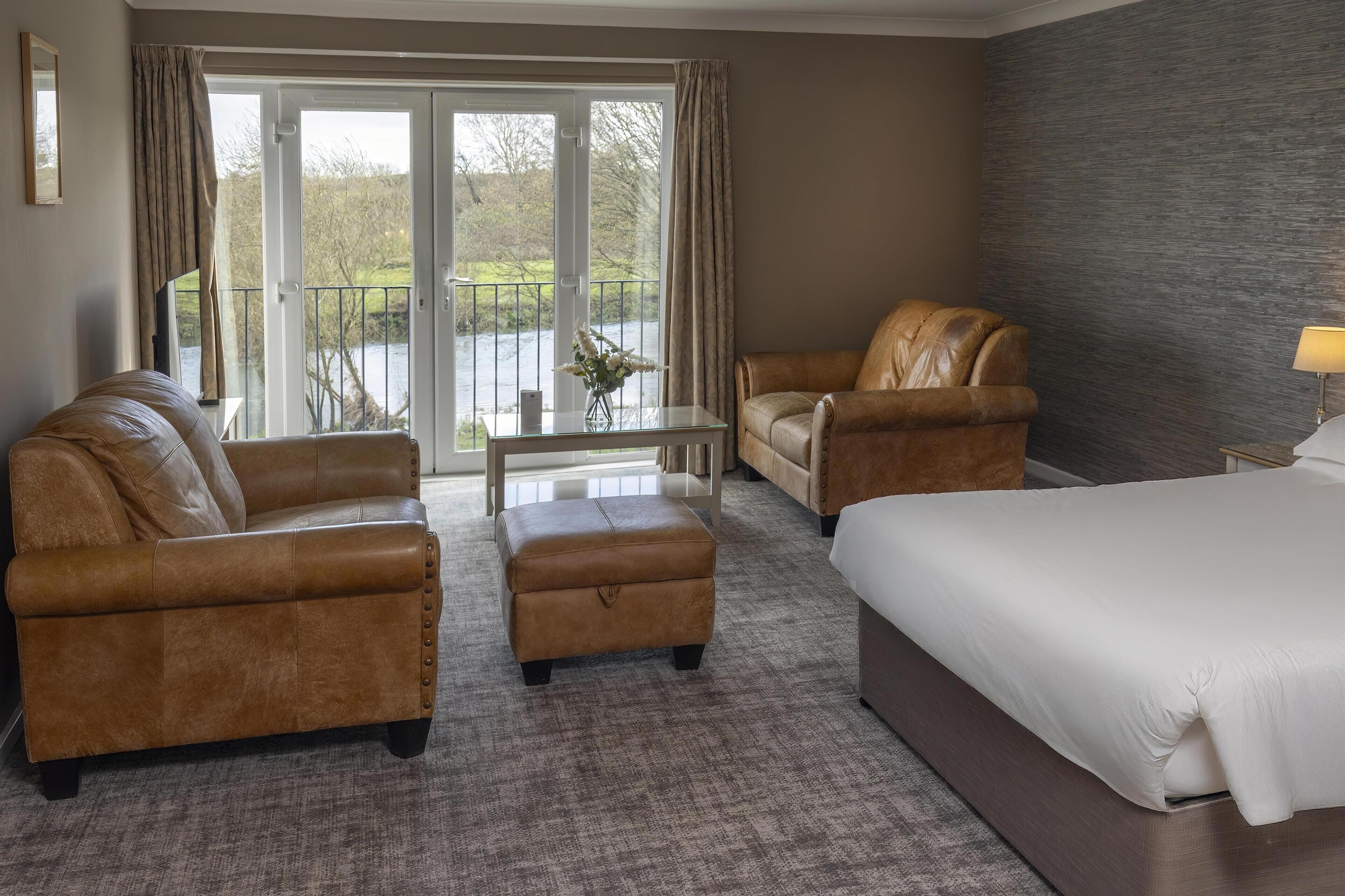 A hotel room with a bed, two leather armchairs, a small ottoman, a table, and a view of a river through large windows.