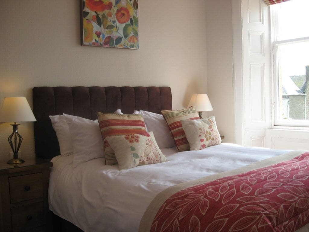 A neatly made bed with white and patterned bedding, two bedside lamps, and a floral painting on the wall.