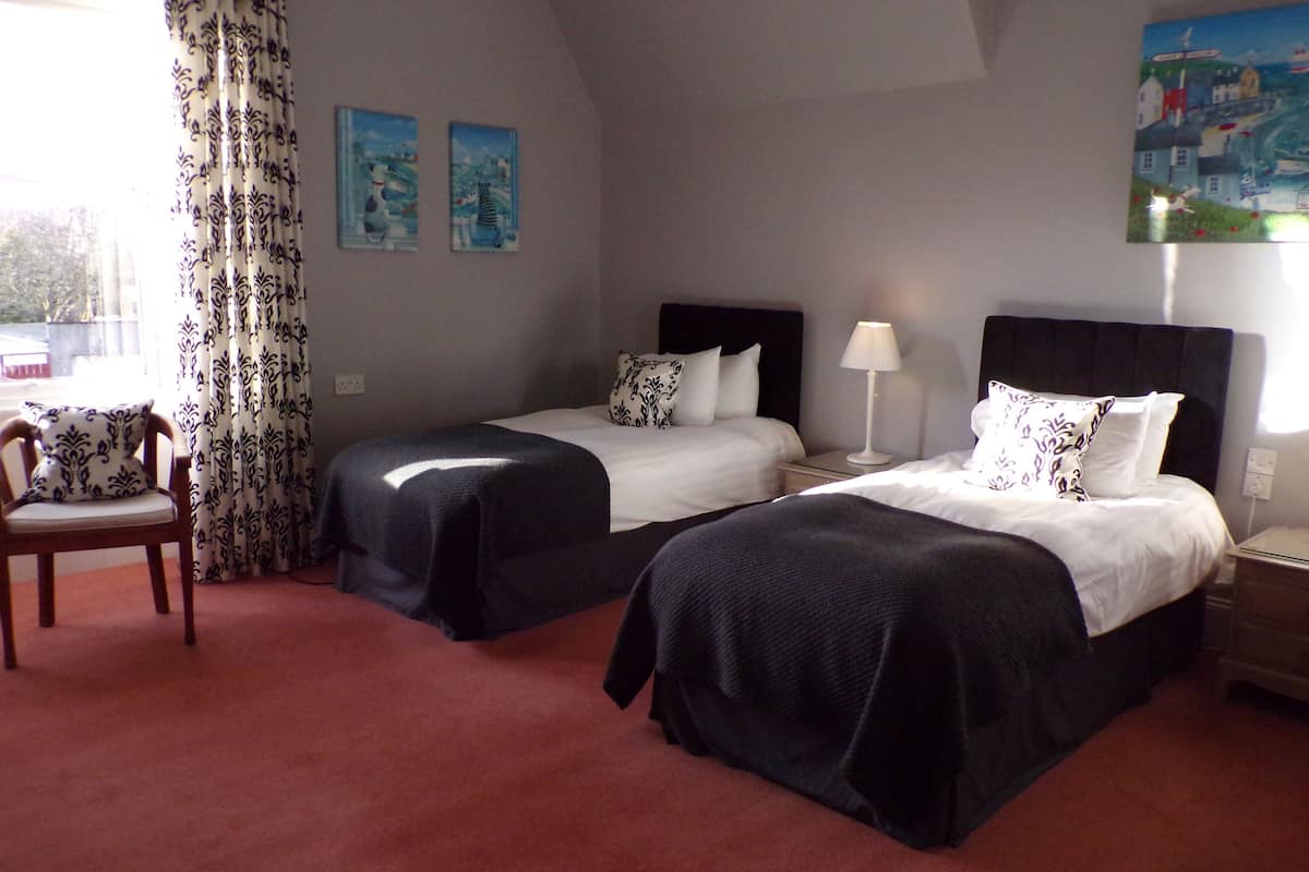 A hotel room with two beds, a chair, a lamp, and two paintings on the wall.