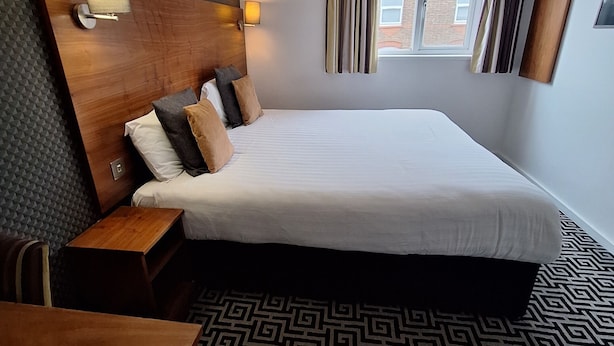 Ein Hotelzimmer mit einem großen Bett, einem hölzernen Kopfteil, einem Nachttisch, einer Pendelleuchte und Blick ins Freie durch ein Fenster mit Vorhängen.