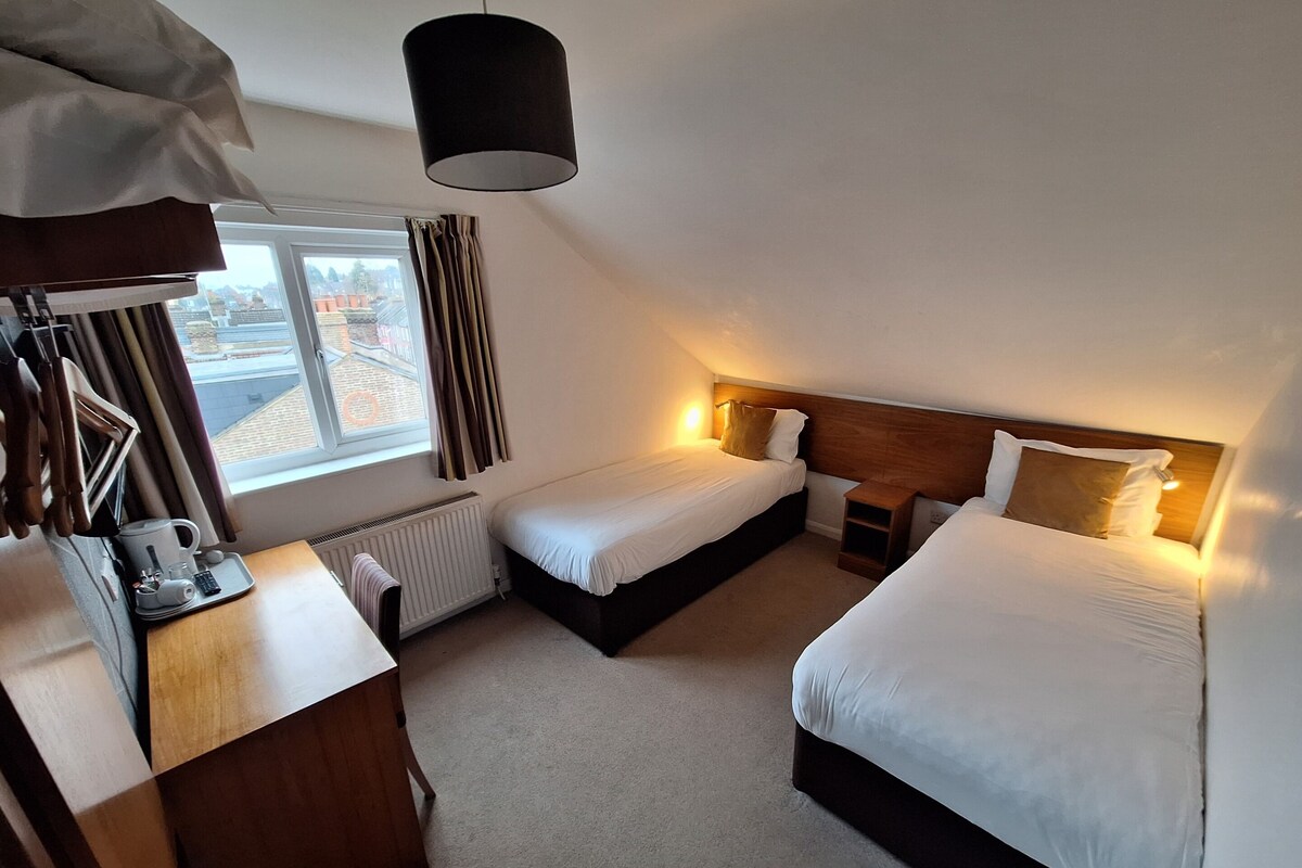 Een hotelkamer met twee bedden, een houten hoofdbord, een raam met gordijnen, een radiator, een nachtkastje met een lamp en een hangende lamp.