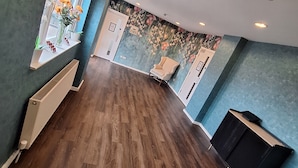 Ein geräumiges Zimmer mit Holzboden, einem großen Fenster mit Blumen und einer Wand mit floralem Wandpapier.