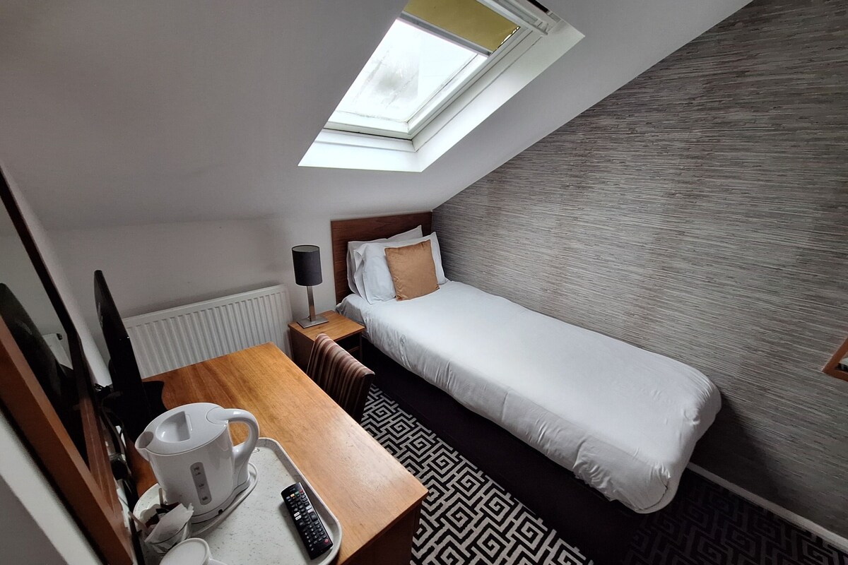 Een compacte hotelkamer met een enkel bed, een klein bureau met een waterkoker, een afstandsbediening en een raam met uitzicht op de lucht.
