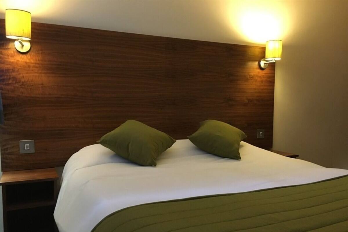 A neatly made bed with a green blanket and two pillows, flanked by wooden headboards with wall-mounted lamps.