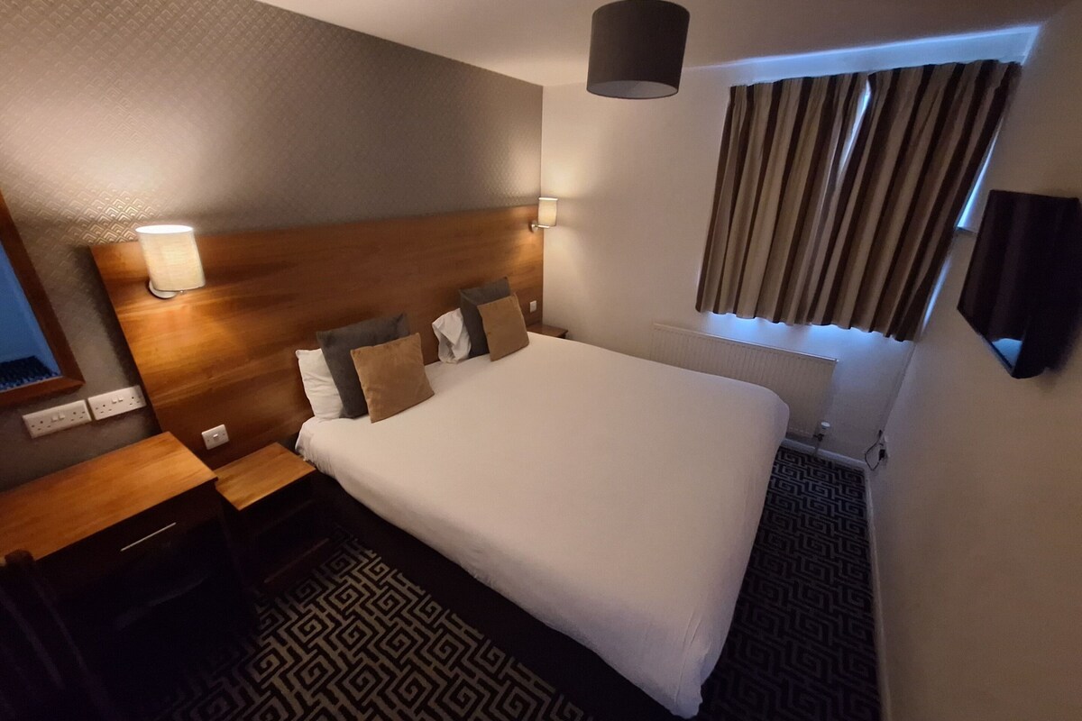 Een hotelkamer met een groot bed, een houten hoofdeinde, een nachtkastje, een lamp, een raam met gordijnen en een kamerbreed tapijt met een patroon.