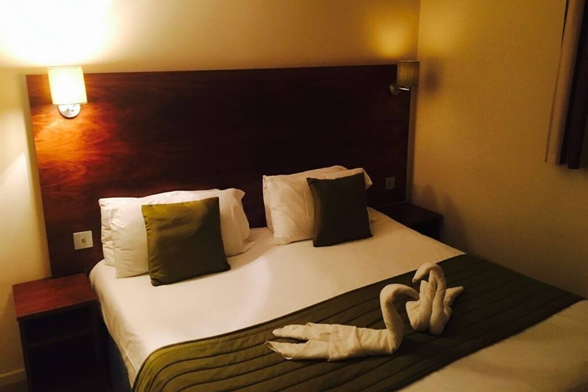 A hotel room with a neatly made bed, a headboard, and a wall-mounted lamp.