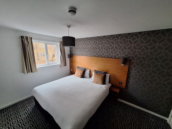 Une chambre d’hôtel moderne avec un grand lit, une tête de lit dotée d’un luminaire, une fenêtre avec des rideaux et une moquette à motifs.