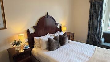 Cama bem arrumada com cabeceira de madeira, duas luminárias de cabeceira, mesa de cabeceira com um vaso e uma xícara, e janela com cortinas.