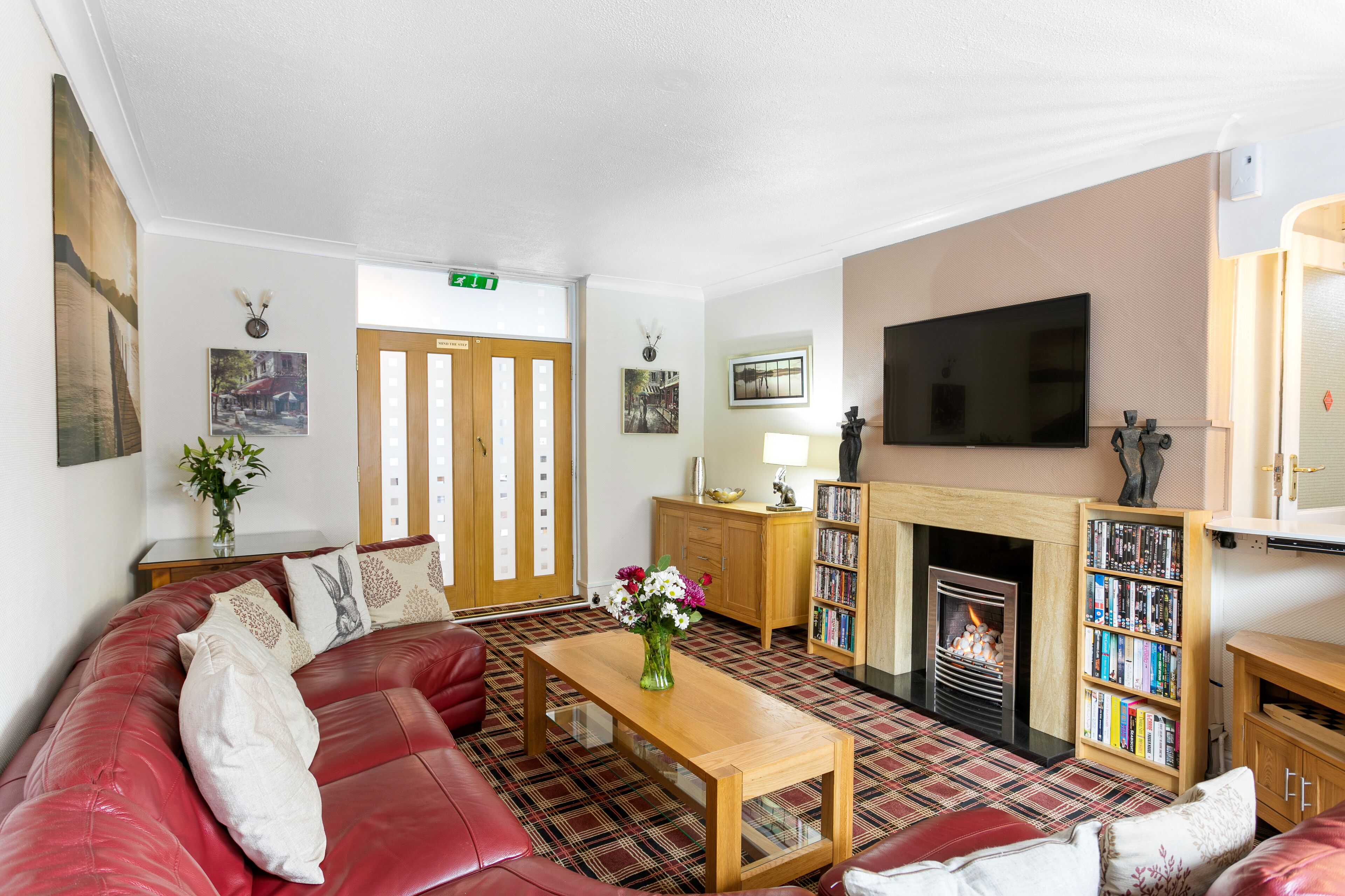 A cozy living room with a red leather sofa, a wooden coffee table, a fireplace, and a bookshelf.