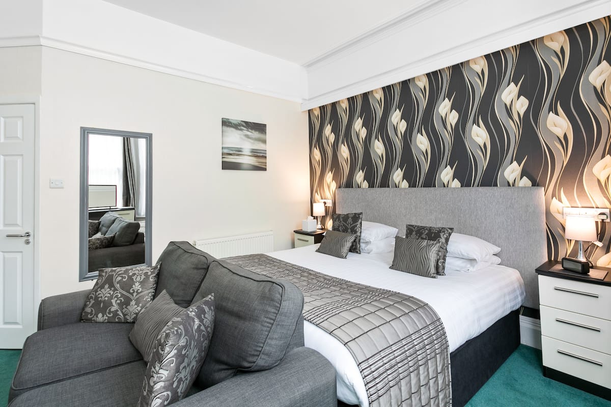 A hotel room with a large bed, a grey sofa, a mirror, a nightstand with a lamp, and a patterned wallpaper.